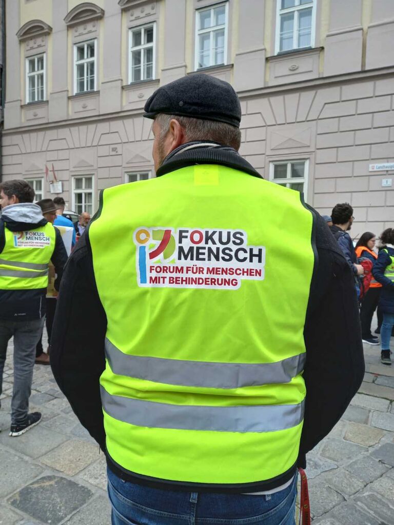 Ein Mann steht im freien und trägt eine Weste mit der Aufschrift "Fokus Mensch, Forum für Menschen mit Behinderung". Im Hintegrund stehen weitere Menschen.
