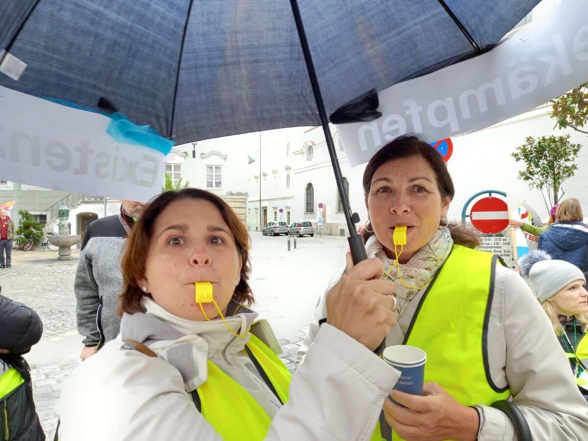 Frauen mit gelber Sicherheitsweste halten Trillerpfeifen im Mund und einen Regenschirm in der Hand, im Hintergrund stehen weitere Personen mit Schildern auf einer Straße.