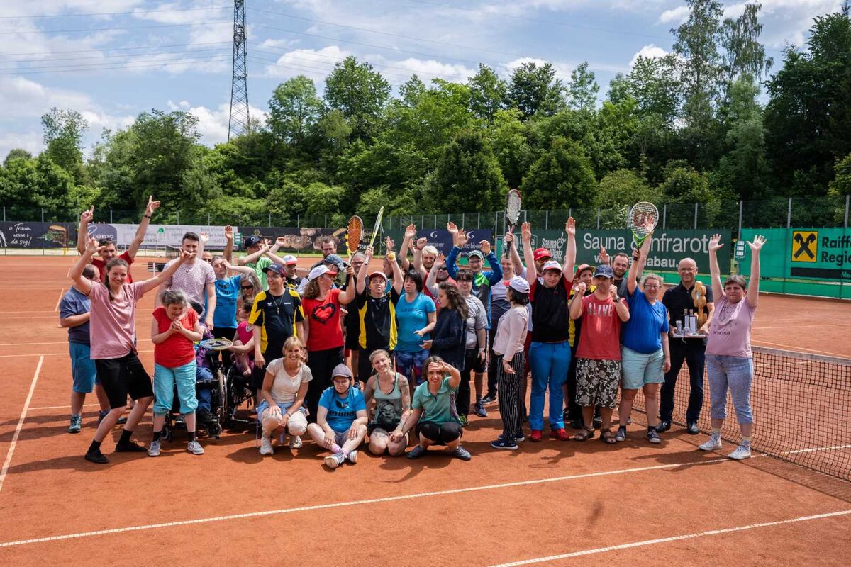 Gruppe von Personen steht auf einem Tennisplatz, mehrere halten Tennisschläger in der Hand und heben die Arme.