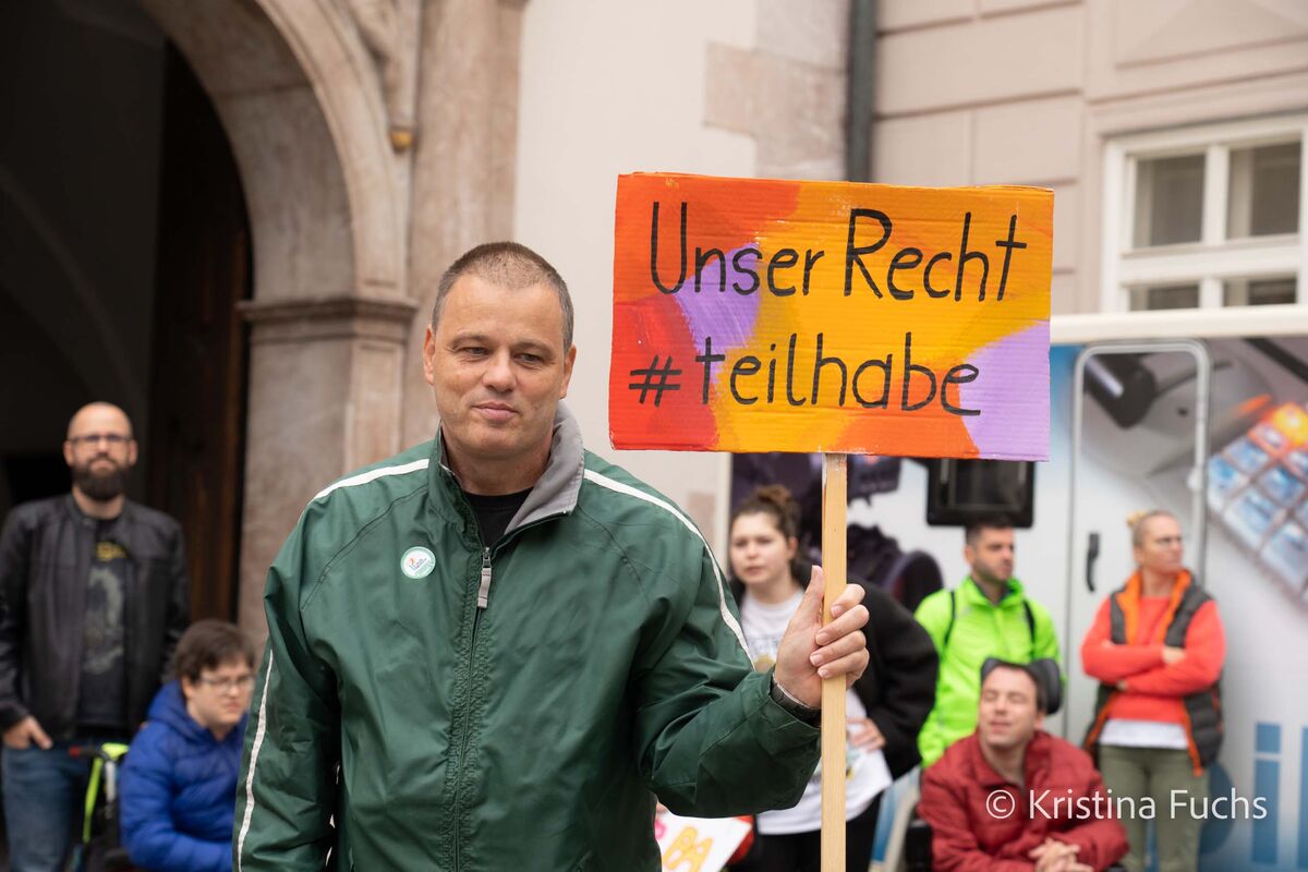 Mann hält bei einer öffentlichen Veranstaltung ein Schild mit der Aufschrift „Unser Recht #teilhabe“, im Hintergrund stehen weitere Personen.