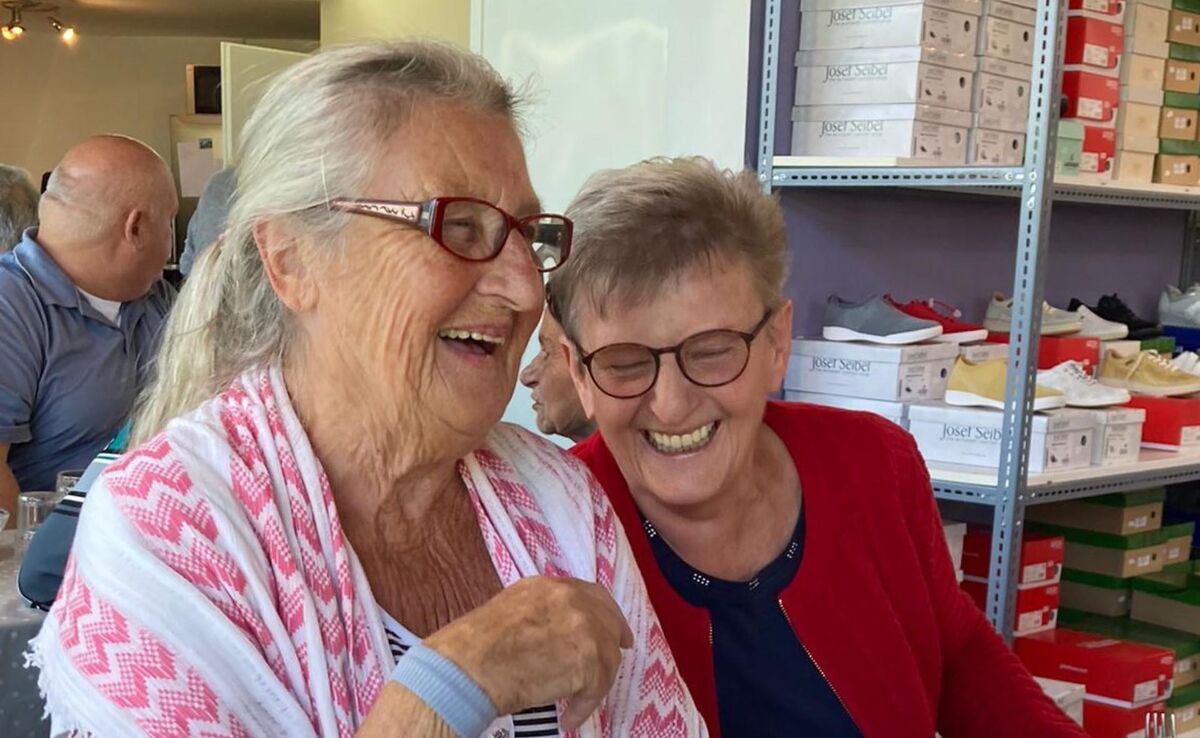 Zwei ältere Frauen mit Brille lachen gemeinsam in einem Raum mit Regalen und Schuhkartons im Hintergrund.