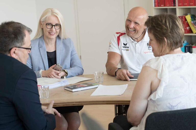 Vier Personen sitzen an einem Tisch in einem Besprechungsraum und führen ein Gespräch mit Unterlagen – Beratung und rechtliche Unterstützung im Team.