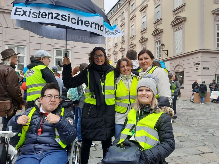 Gruppe von Personen mit gelben Warnwesten steht auf einem Platz, eine Person sitzt im Rollstuhl, im Hintergrund ist ein Regenschirm mit der Aufschrift „Existenzsichernde Arbeit“ zu sehen.