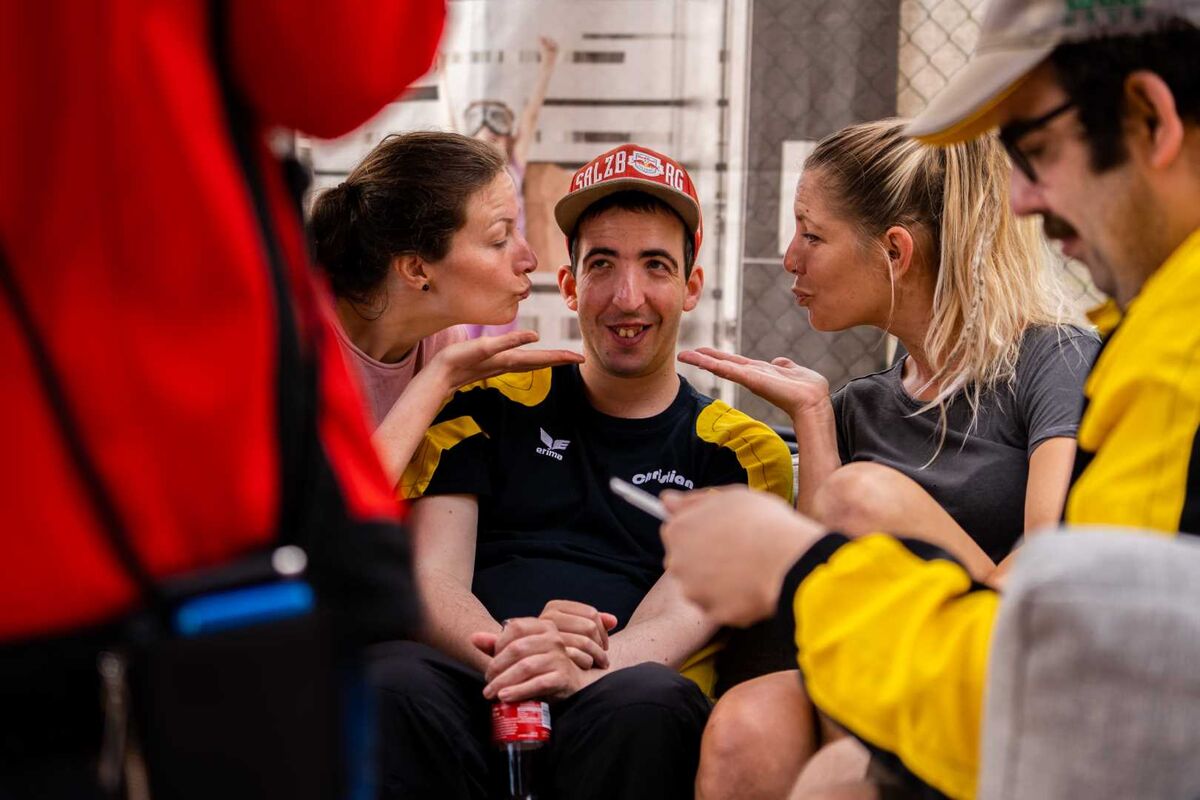 Ein Mann sitzt lächelnd in Sportkleidung, zwei Frauen beugen sich von links und rechts zu ihm und sprechen gestikulierend; im Vordergrund ist eine weitere Person unscharf zu sehen.