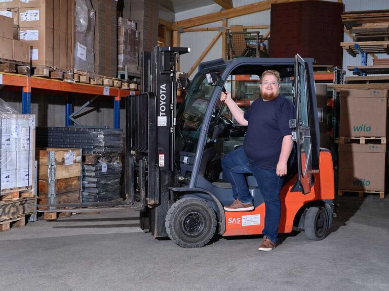 Person fährt einen Gabelstapler in einer Lagerhalle zwischen Regalen mit Waren – betriebliche Tätigkeit im Arbeitsalltag.