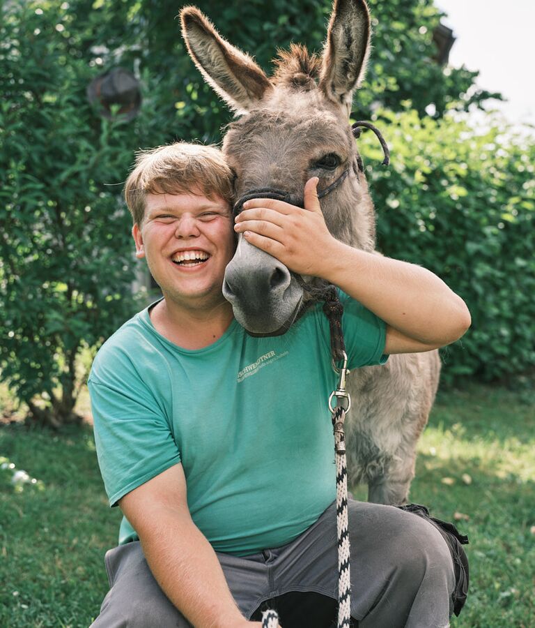 Ein junger Mann steht im Freien neben einem Esel und hält das Tier am Halfter.