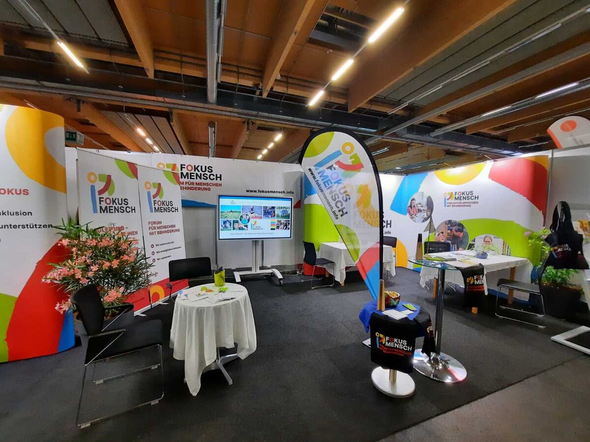 Ein Bild eines Informations- und Messestandes mit Bannern, Roll ups und Beach flags von Fokus Mensch in einer Messehalle.
