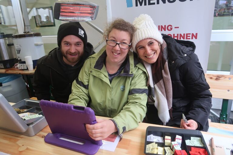 Drei Erwachsene sitzen an einem Tisch, schauen auf ein Tablet und lächeln in die Kamera.