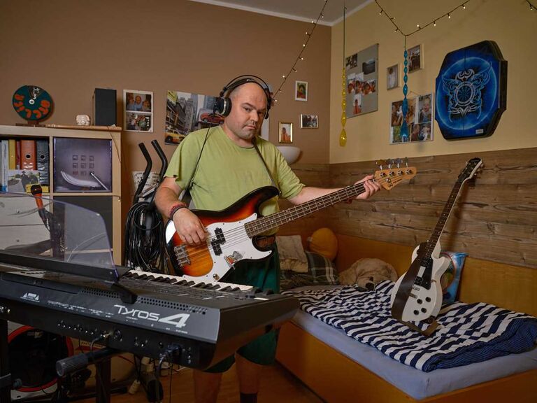 Ein Mann sitzt in seinem Zimmer mit Kopfhörern und spielt E-Bass, vor ihm stehen Keyboard und weitere Instrumente – individuelles Wohnen mit Raum für persönliche Interessen.