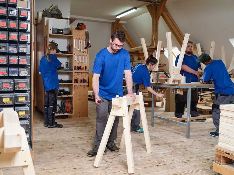 Mehrere Personen arbeiten in einer Holzwerkstatt an Werkbänken und fertigen Werkstücke – praktische Tätigkeit in strukturierter Arbeitsumgebung.
