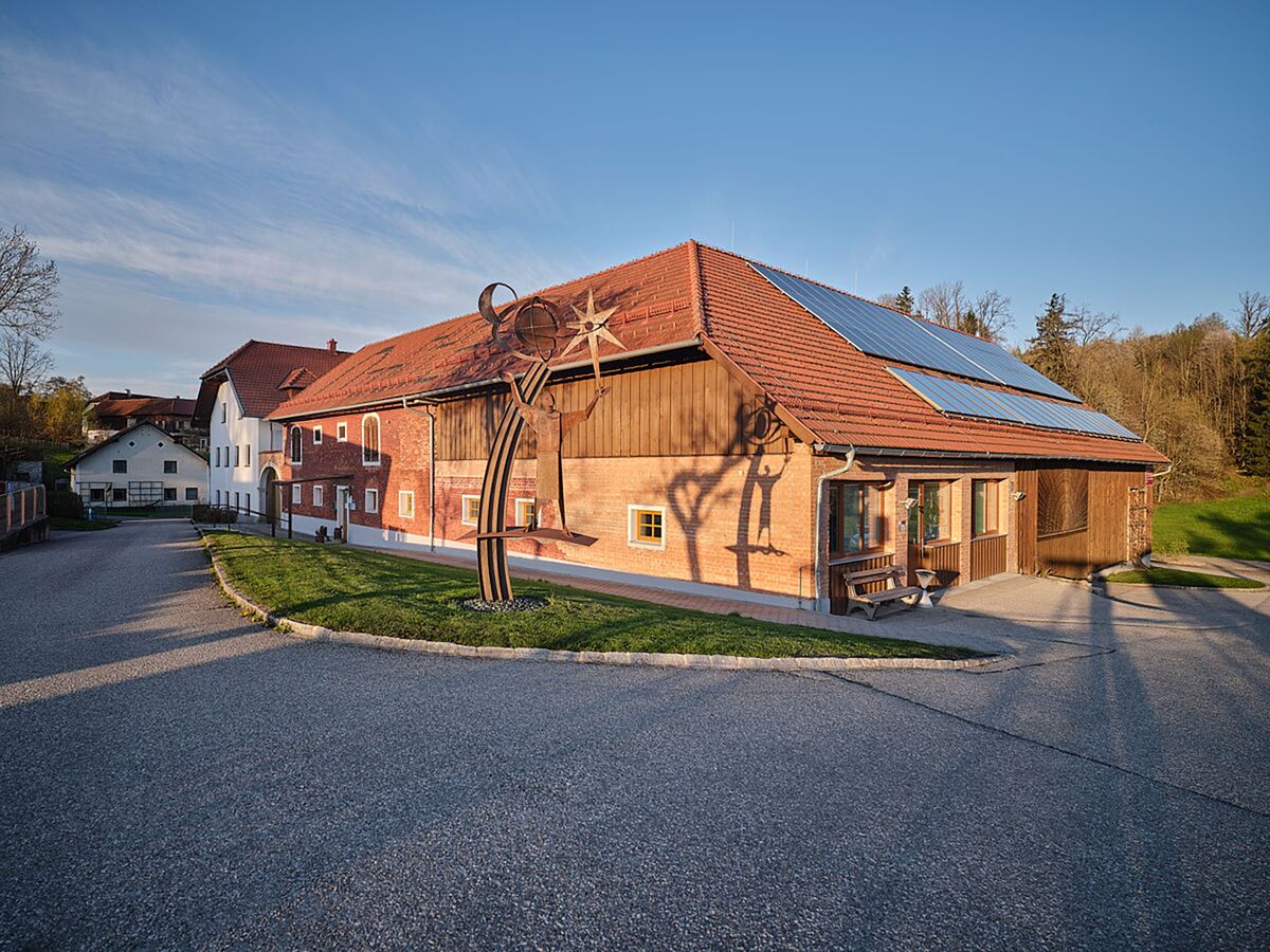 Holzgebäude mit Satteldach und Solarpaneelen, davor ein gepflasterter Vorplatz.