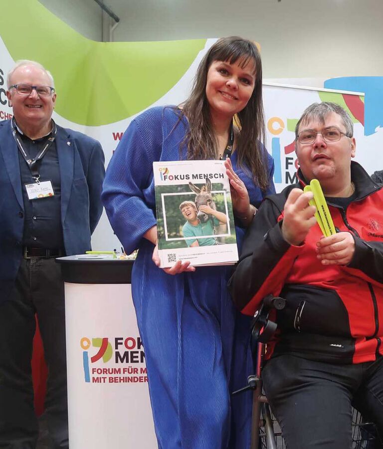 Zwei Frauen und ein Mann präsentieren gemeinsam einen Flyer von Fokus Mensch auf einer Veranstaltung.