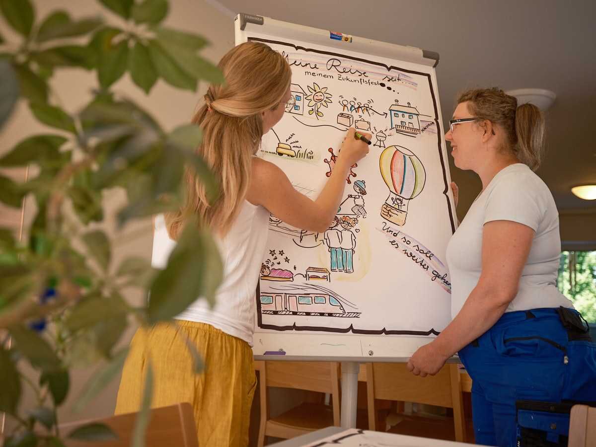 Eine Frau erklärt Inhalte auf einem Whiteboard, eine Person hört aufmerksam zu.