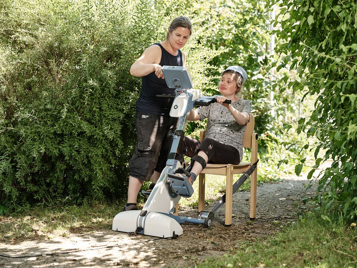 Zwei Frauen befinden sich im Freien neben einem Trainingsgerät, eine Person sitzt auf dem Gerät, die andere steht daneben und unterstützt.