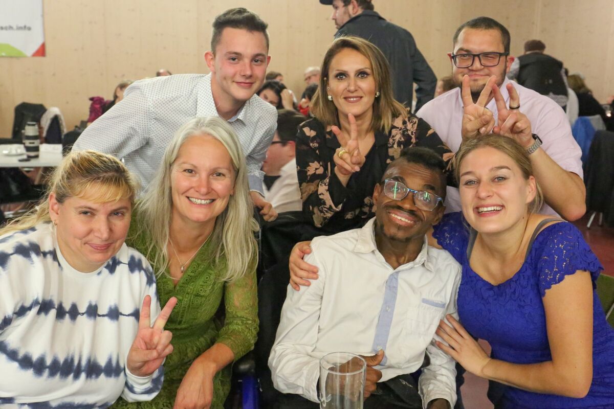 Mehrere Erwachsene sitzen eng beieinander und lächeln in die Kamera, eine Person legt einem Mann den Arm um die Schulter.