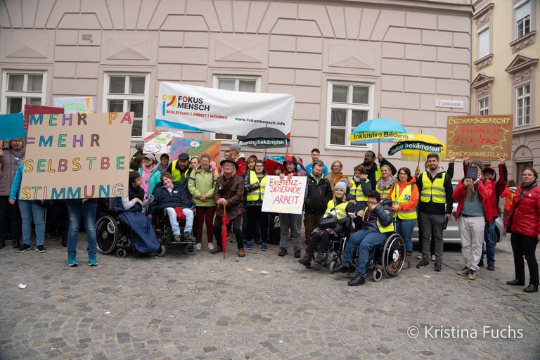 Gruppe von Personen steht auf einem Platz vor einem Gebäude, einige halten Banner und Schilder, mehrere Personen nutzen Rollstühle.