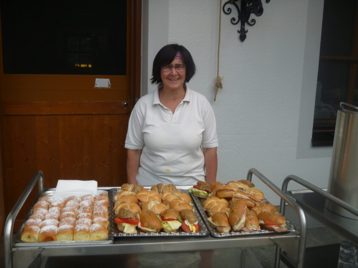 Eine Frau steht hinter einem Servierwagen, auf dem Semmeln und Buchteln angerichtet sind.