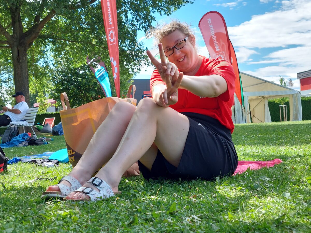 Teilnehmerin sitzt in der Wiese, lacht, macht Peace-Zeichen