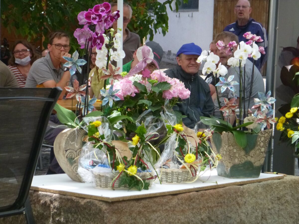 im Vordergrund stehen mehrere Blumenstöcke, im Hintergrund sihet man einige Gäste sitzen