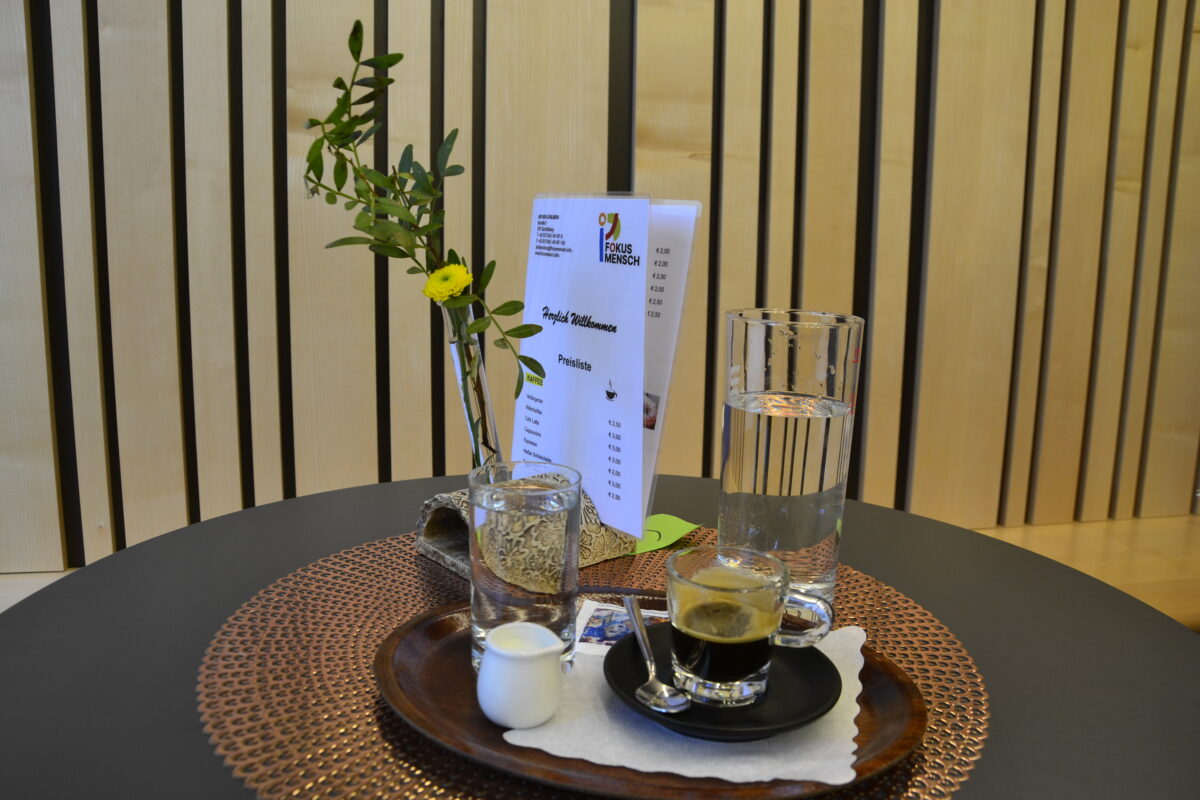 Ein Espresso und ein großes Glas Wasser sowie eine Blumenvase, dahinter eine Speisekarte, stehen auf einem Kaffeetisch, 