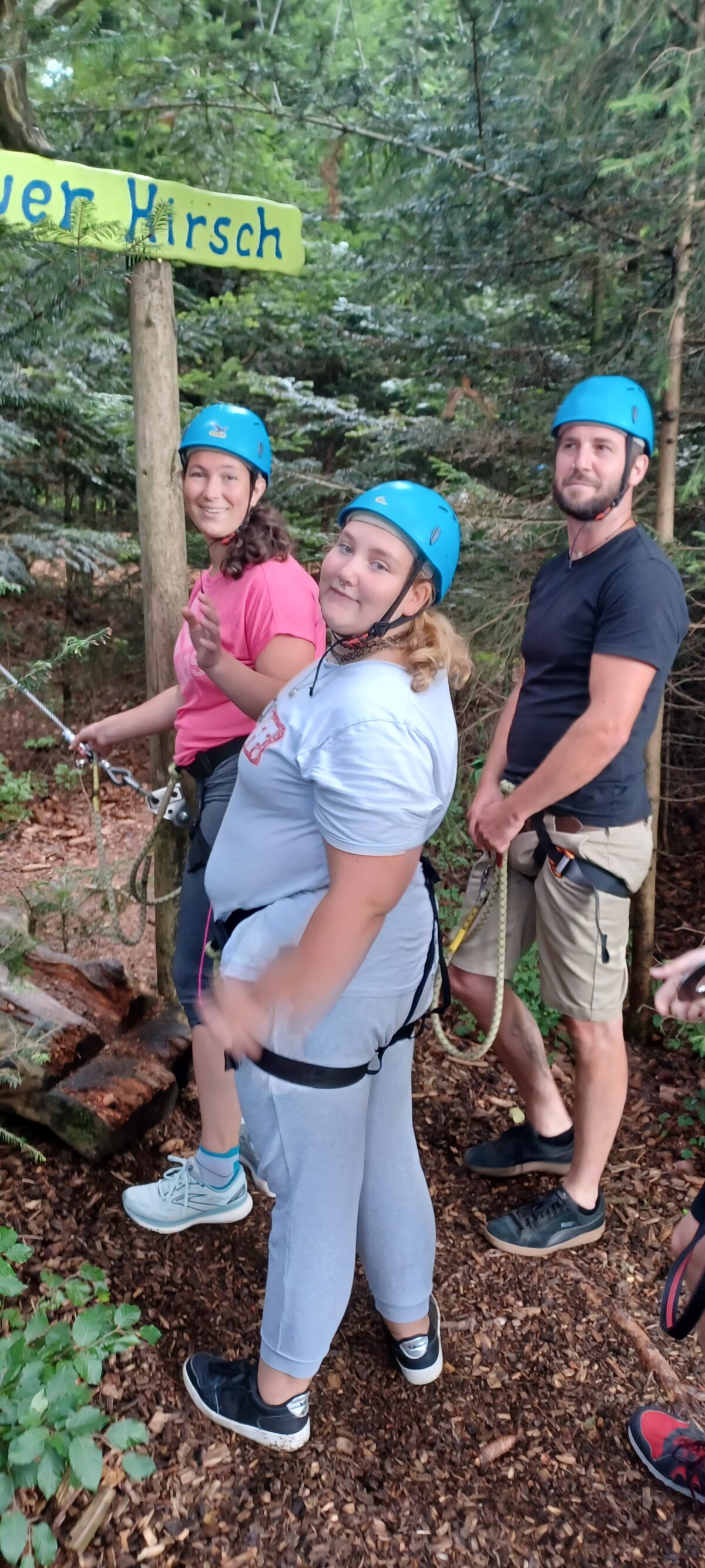 zwei junge Frauen und ein Mann mit Kletterhelmen und Klettergurt am Einstieg eines Hochseilklettergartens