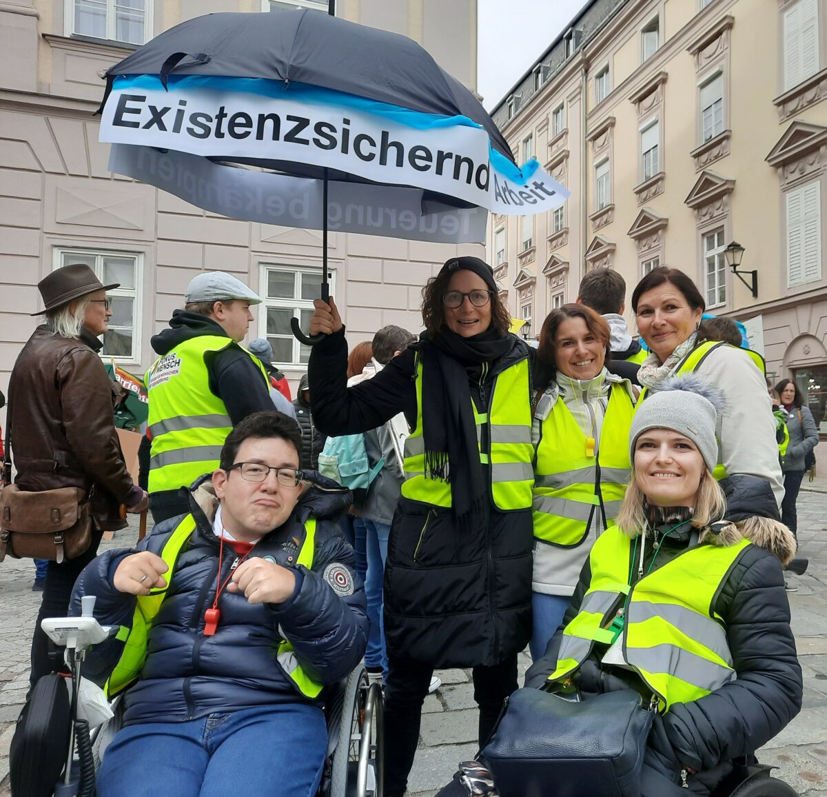 Gruppe von Demonstrierenden in Warnwesten und mit Regenschirm mit der Botschaft "Existenzsichernde Arbeit"