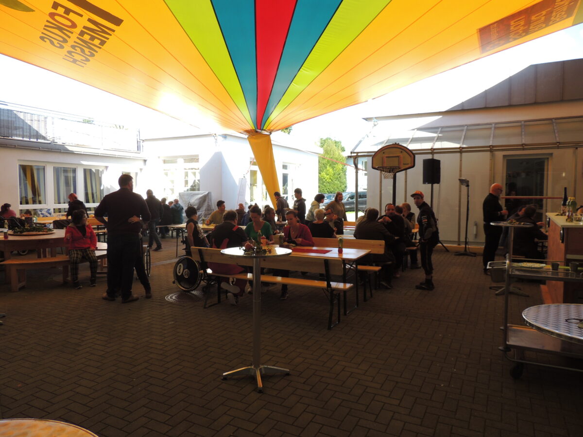 gelbe Plane über dem Innenhof, wo Biertische und Bänke aufgestellt sind, auf denen Menschen sitzen und plaudern