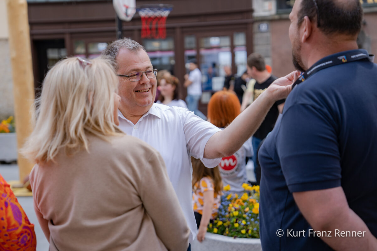 drei Personen, in der Mitte ein fröhlicher Mann, der wohin deutet
