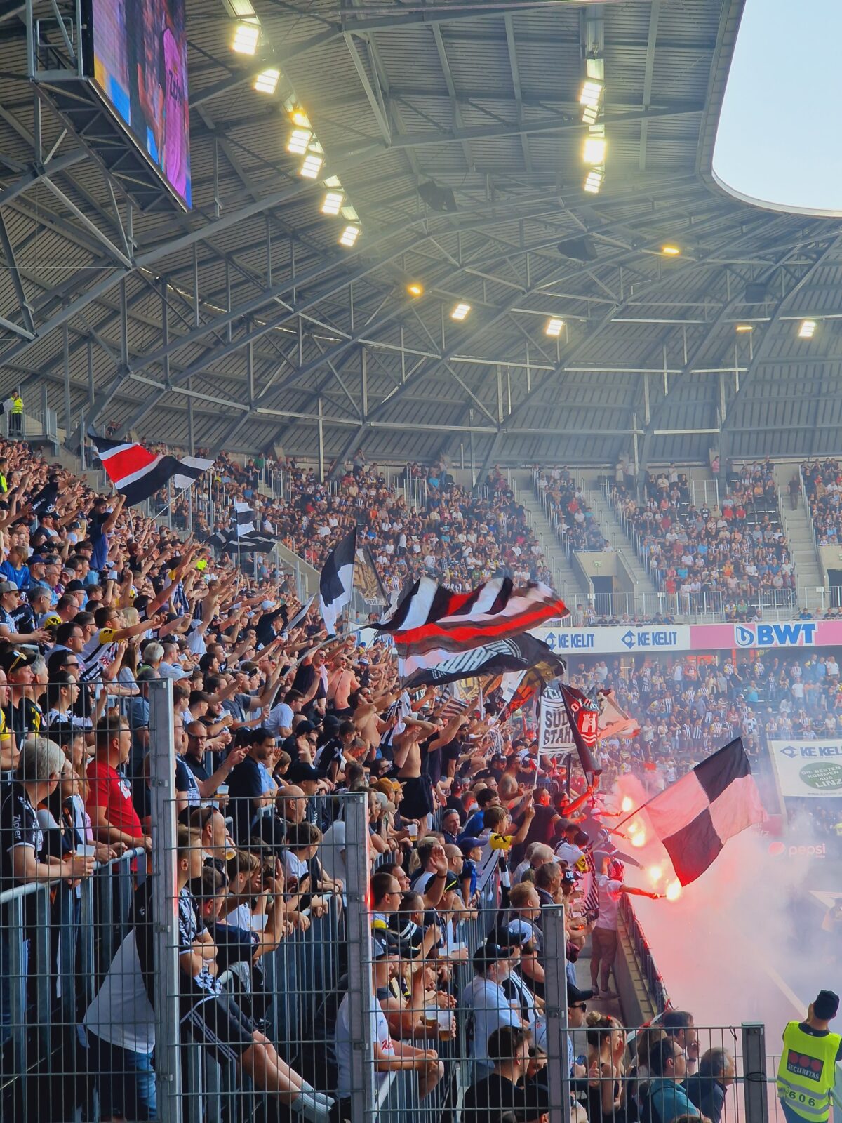 Blick in die Menschenmenge auf der Tribüne, Fahnen werden geschwenkt und Feuersticks abgefackelt