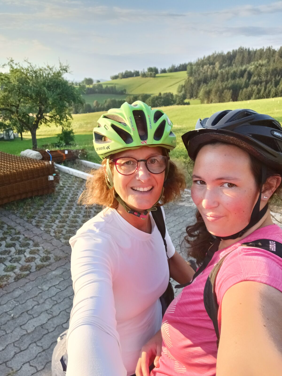 Selfie mit zwei Frauen, die einen Fahrradhelm tragen, im Hintergrund ist eine Landschaft mit Wiesen und Wälder