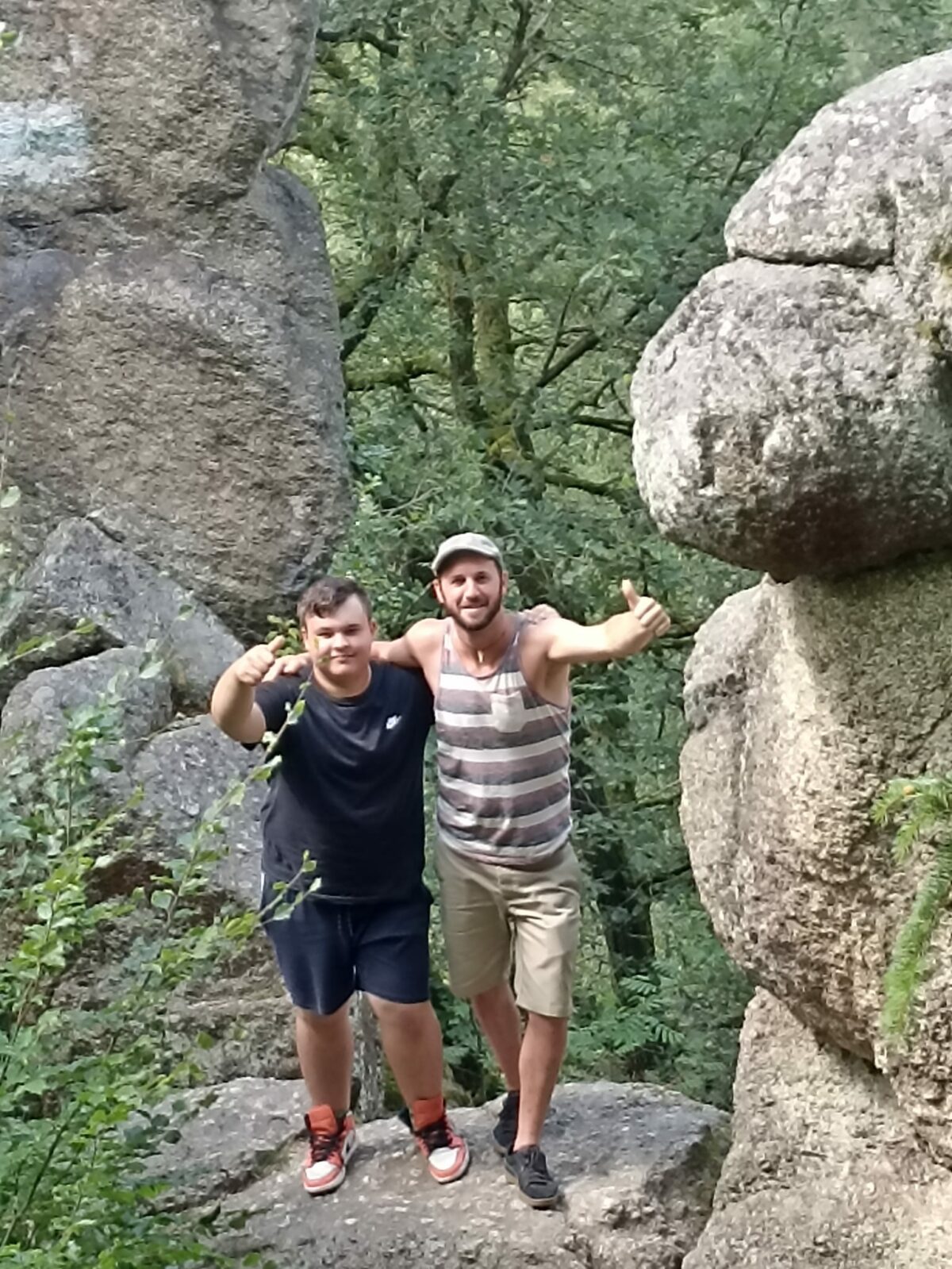 zwei Männer im Wald zwischen großen Steinen beim Wandern, lachen in die Kamera, Daumen nach oben