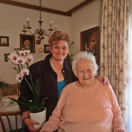 eine sehr alte Frau und eine Frau mit Orchideen-Blumenstock lachen in die Kamera