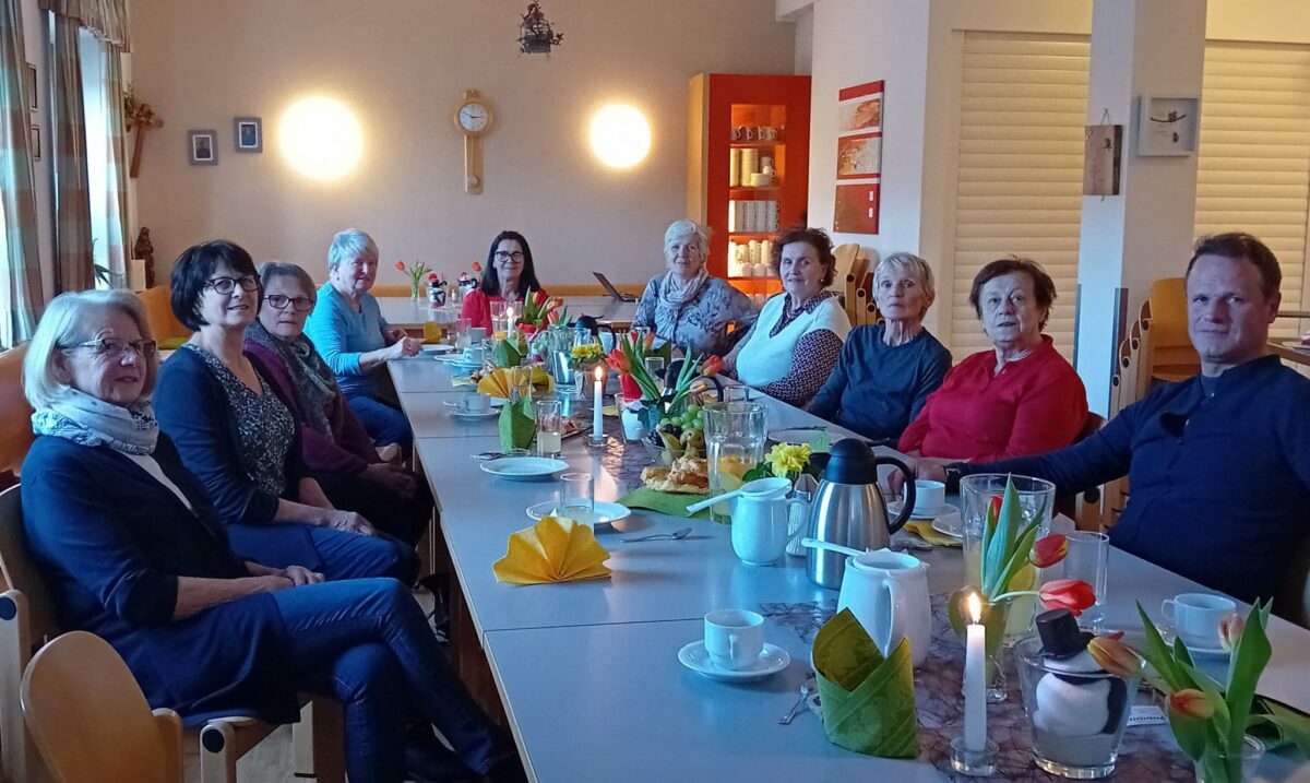 neun ältere Frauen und ein Mann sitzen bei einem mit Kaffee und Nachspeise gedeckten Tisch
