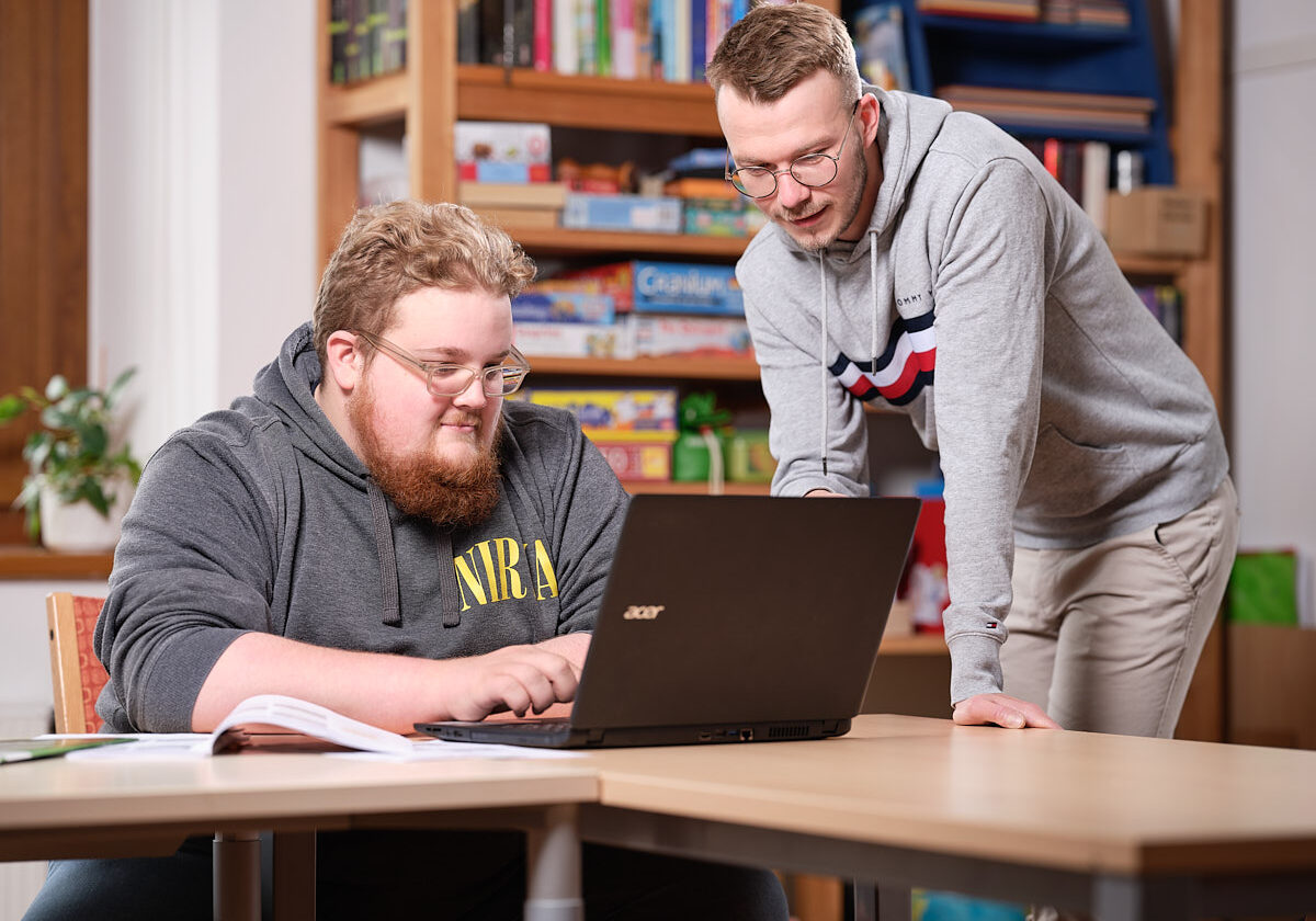 junger Mann sitzt bei einem Laptop, ein anderer junger Mann beugt sich zu ihm und deutet auf den Bildschirm