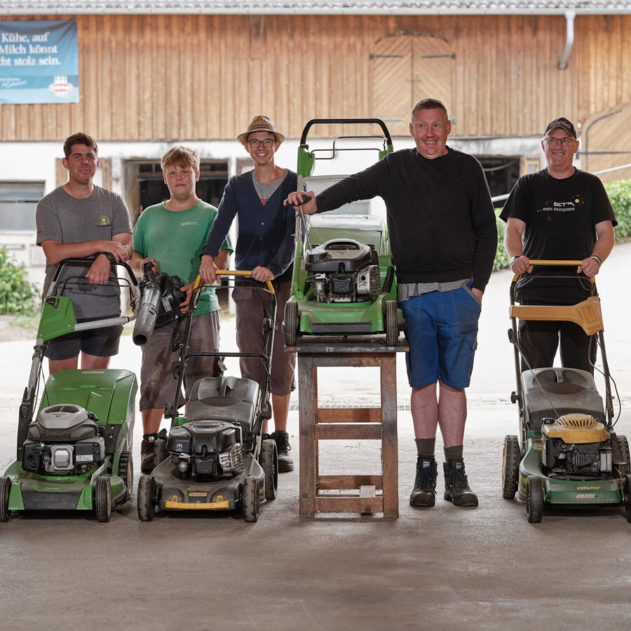fünf fröhliche Männer mit Blick zur Kamera stehen bei Rasenmäher-Geräten