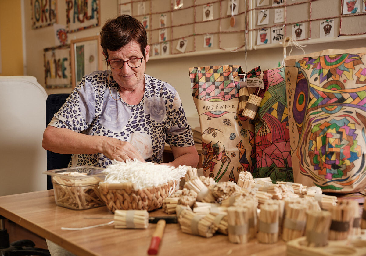 ältere Frau arbeitet bei Tisch mit kleinen Holzstücken, bunt bemalte Papiersäcke stehen daneben