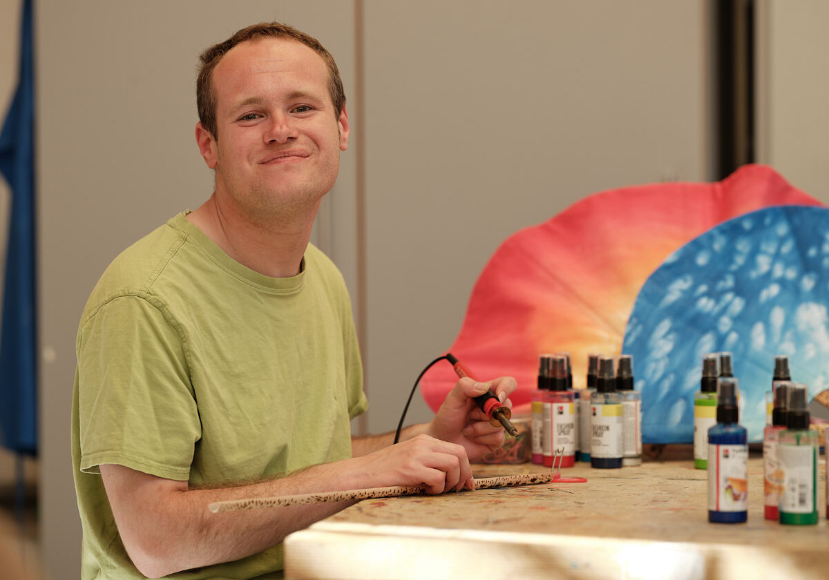 junger Mann bei mit Brennkolben, bunte Farbfläschchen stehen am Tisch, zwei Sauna-Fächer im Hintergrund