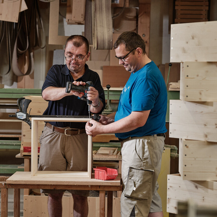 Mann mit Akku-Bohrer fertigt Holzkisten an und wird von einem zweiten Mann dabei unterstützt
