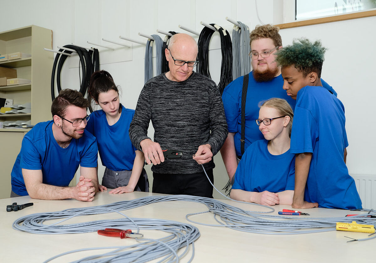 fünf junge Menschen stehen bei einem Betreuer, der ihnen einen Arbeitsschritt bei Kabeln zeigt