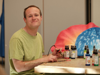 junger Mann bei mit Brennkolben, bunte Farbfläschchen stehen am Tisch, zwei Sauna-Fächer im Hintergrund