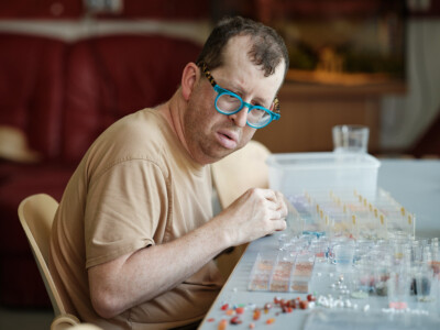 Mann mit blauer Brille sortiert bei Tisch kleine bunte Perlen