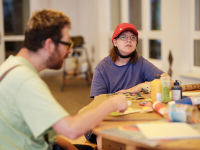 junge Frau mit roter Kappe sitzt bei Tisch mit Kreativ-Werkstücken und blickt gedankenverloren nach oben