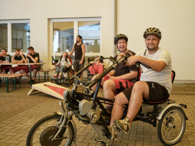 Gruppe von Menschen verbringt entspannte Zeit im Innenhof des Feichtlguts, im Vordergrund: zwei Männer auf einem Therapie-Tandem